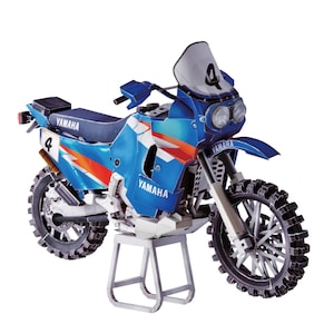May include: A blue and white Yamaha motorcycle model with orange detailing. The model displays the number 4 on the side and windshield. The tyres are black and the motorcycle is on a silver stand.