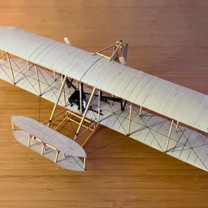 May include: A model airplane with a biplane design, featuring a white fabric-covered wing and tail. The wooden frame and struts are visible, and the plane rests on a wooden surface. The model includes a pontoon for water landings.