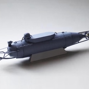 May include: A model of a submarine, painted in a dark gray color. The submarine has a cylindrical body with a raised section on top and a framework at the front and rear. The model is displayed on a white surface.