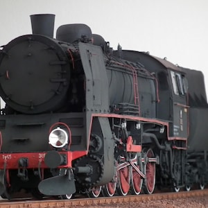 May include: A detailed model of a black steam locomotive with red accents on the wheels and trim. The model features two large headlights and a coal tender. The locomotive is set on a brown track.