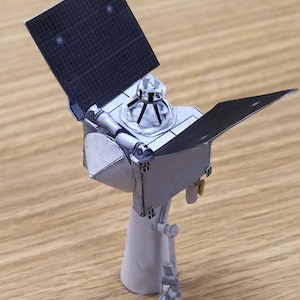 May include: A paper model of a spacecraft, predominantly light grey in colour. It features a central body, extended black solar panels, and a cylindrical base. The model has detailed elements, including a grid pattern on the panels.