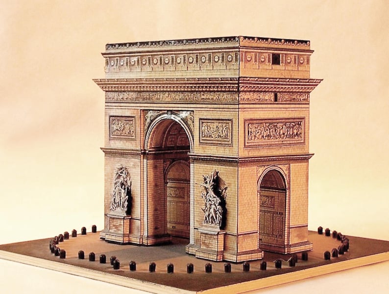 May include: A detailed model of the Arc de Triomphe, featuring intricate architectural details. The model is primarily beige and brown, with sculpted figures and archways. It sits on a rectangular base with small, dark posts around the perimeter.