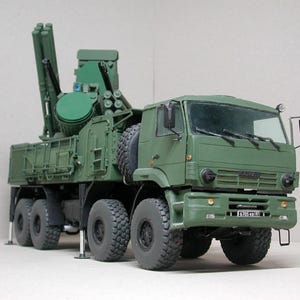 May include: A detailed model of a military truck in olive green. The truck features multiple wheels, a cab, and a complex structure on the back with missile launchers. The model is set against a plain background.