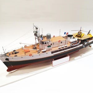 Puede incluir: Maqueta detallada de un barco con una plataforma para helicópteros. El barco presenta una cubierta de madera, un casco blanco y negro y varias antenas y equipos. Un pequeño helicóptero amarillo está en la plataforma. Se ven banderas francesas.