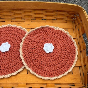 May include: Two round, crocheted coasters featuring a rust-colored center, a scalloped beige border, and a small white flower-shaped accent in the middle of each piece. The coasters are placed inside a woven wooden basket with a light brown finish.