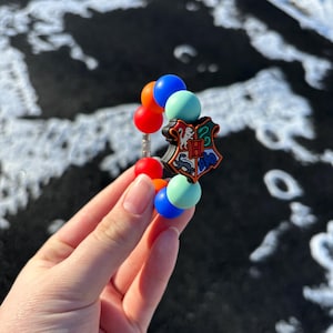 May include: A colorful bracelet featuring a Harry Potter Hogwarts crest charm and several round beads in red, orange, blue, and green. The bracelet is held against a snowy background.