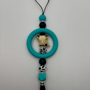 May include: A teal-colored teething ring with a Highland cow charm, black and white cow print beads, and a black tassel. The charm is brown and tan. The top has a black cord and a black clasp.