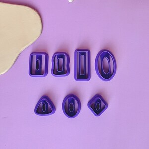 Puede incluir: Un juego de ocho cortadores de galletas de plástico morado en varias formas, incluyendo círculos, cuadrados, rectángulos y diamantes. Los cortadores están dispuestos sobre una superficie de color morado claro.