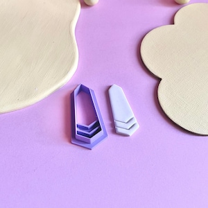 Könnte beinhalten: Zwei violette Plastik-Ausstecher, einer in Chevronform und der andere in Rechteckform mit einem Chevron-Muster. Die Ausstecher befinden sich auf einer hellvioletten Oberfläche.