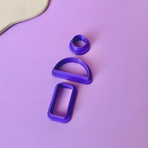Puede incluir: Tres cortadores de galletas de plástico morado en forma de círculo, semicírculo y rectángulo.
