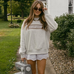Puede incluir: Una sudadera gris claro de cuello redondo con el texto marrón "HOT GIRLS HAVE GRANNY HOBBIES" en un marco rectangular, combinada con pantalones cortos blancos con ribete de encaje, botas estilo western color crema y una máquina de coser gris.