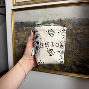May include: A beige fabric drink sleeve with a black paisley pattern and the word "MOTHER" in brown letters. The sleeve has a tag that says "COPEN CO." and is holding a clear glass.