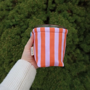 May include: A small, cylindrical container with a glass top and a fabric exterior. The fabric features vertical orange and light purple stripes. The container is held in a hand against a backdrop of green foliage. The text "COPEN CO." is visible on the side.