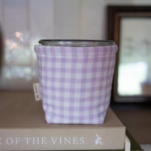 May include: A cylindrical fabric sleeve featuring a light purple and white gingham check pattern. The sleeve has a small white fabric tag on the side with the text COPIN CO. printed in black. The top edge is finished with a clean stitched hem.