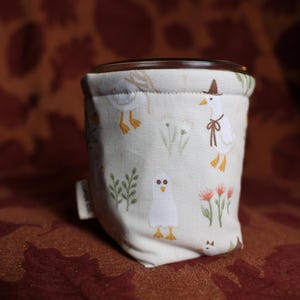 May include: A cylindrical fabric container with a cream-colored background featuring a repeating pattern of white geese, some wearing brown hats and scarves, interspersed with green foliage and orange flowers. A small white tag reads Coper Co.