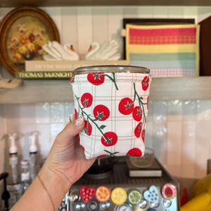 May include: A cylindrical fabric cup sleeve featuring a white background with a light beige grid pattern and a repeating print of red tomatoes on green vines. The sleeve has a finished top edge and a small white fabric tag on the side.