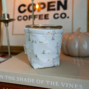 May include: A fabric cup sleeve featuring a repeating pattern of white geese with pink bows and small pink flowers on a light gray background. A small white fabric tag on the side of the sleeve displays the text COPEN CO. in black lettering.