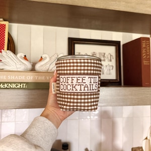 May include: A cylindrical glass tumbler featuring a brown and white gingham fabric sleeve. The sleeve displays the embroidered text COFFEE TIL COCKTAILS in a block font and includes a small white fabric tag labelled COPEN CO. on the side.