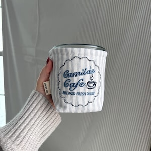 Puede incluir: Una funda para taza de café a rayas blancas y grises con el texto bordado "Camila's Cafe BREWED FRESH DAILY" en azul. La funda tiene un borde festoneado y una pequeña etiqueta que dice "COFFY CO."