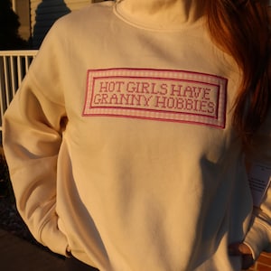 May include: A cream-coloured sweatshirt with a high neck, featuring the embroidered text "HOT GIRLS HAVE GRANNY HOBBIES" in a pink rectangular frame. The text is in a cross-stitch style. The sweatshirt is a neutral colour.