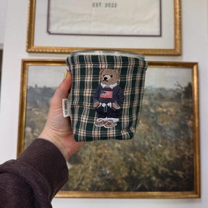 May include: A green and beige plaid cup cozy with an embroidered bear wearing a navy blue jacket and an American flag. The cozy has a small white tag that says "COPE CO."