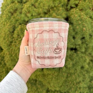 May include: A light pink and white gingham fabric cup sleeve featuring an embroidered scalloped design with the text Ashley's Cafe and Brewed Fresh Daily, including a small coffee cup icon and a side tag labeled COPEN CO.
