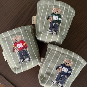 May include: Three small fabric storage baskets in olive green with white vertical stripes. Each basket features an embroidered teddy bear wearing a sweater with the number 00 and holding a football. A small white tag reads COPEN CO.