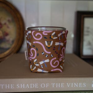 May include: A brown fabric bowl cozy featuring a repeating pattern of pink earthworms, yellow and black bees, blue dragonflies, and small red ladybugs. The cylindrical container has a visible top rim and a small white fabric tag on the side.