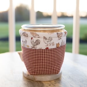 May include: A glass jar with a lid is encased in a fabric cozy. The top portion of the cozy features a white background with chicken illustrations. The bottom portion is a red and white checkered pattern. The jar sits on a white marble coaster.