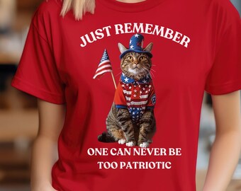 May include: Red t-shirt featuring a patriotic cat wearing a hat and holding an American flag. The shirt has the text "JUST REMEMBER ONE CAN NEVER BE TOO PATRIOTIC" in white. The cat is dressed in red, white, and blue.