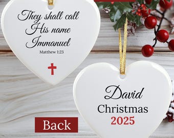 May include: White heart-shaped Christmas ornament with gold ribbon. The front has the text "They shall call His name Immanuel" and a red cross. The back says "David Christmas 2025".