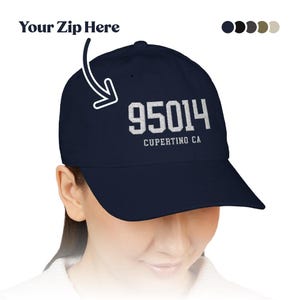 Puede incluir: Una gorra de béisbol azul marino con el texto bordado en blanco 95014 y CUPERTINO CA en la parte delantera. Un gráfico de flecha señala los números con el texto Your Zip Here encima. Cinco pequeños círculos de colores aparecen en la esquina.