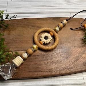May include: A wooden car air freshener with a daisy design. The air freshener features a wooden ring with a daisy in the centre, surrounded by small orange accents. It has a string for hanging and decorative wooden beads.