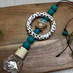 Könnte beinhalten: Türkisfarbener und brauner Auto-Lufterfrischer mit einem Kuh-Anhänger. Der Anhänger trägt eine blaue Baseballkappe. Der Lufterfrischer ist an einer braunen Schnur befestigt.