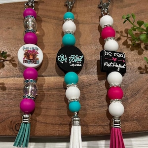 Peut inclure: Trois porte-clés perlés avec des fermoirs argentés. Chaque porte-clés présente un charme différent avec le texte : "Just a lil Moody", "Be Kind...of a Bitch" et "be real Not Perfect". Les porte-clés ont des perles turquoise, blanches, roses et transparentes.