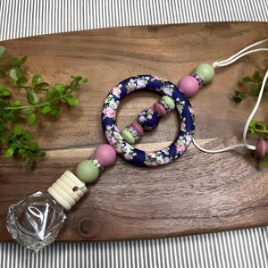 Op de afbeelding: Een autoluchtverfrisser met een bloemenprint en een kralenketting. De ketting heeft roze, groene en witte kralen. De luchtverfrisser is bevestigd aan een heldere glazen fles met een houten dop.