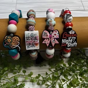 May include: Four colorful bracelets with charms. The charms are a leopard print stethoscope, a coffee cup with the words "Coffee Scrubs", a pink bow with a coffee cup and a red cross, and a crown with the words "Queen of the Night Shift".