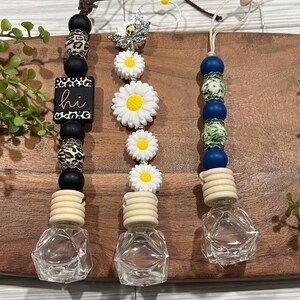 May include: Three car air fresheners with different designs. The first is black and white with a leopard print and the word "hi" on a wooden tag. The second is white with a daisy design. The third is blue and green with a floral design. All three have a clear glass bottle with a wooden top.
