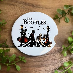 May include: White ceramic car coaster with a Halloween-themed design. The coaster features the text "The Boo-tles" above a graphic of costumed figures crossing a crosswalk. A witch, a skeleton, and a mummy are depicted, along with bats and a traffic light.