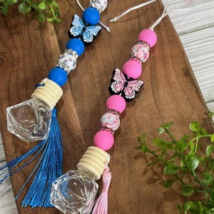 Pode incluir: Dois aromatizadores de carro com pingentes de borboleta. Um é azul com uma borboleta azul e o outro é rosa com uma borboleta rosa. Ambos têm uma garrafa de vidro transparente com uma rolha de madeira e uma borla.