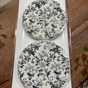 May include: Two white ceramic car coasters with a black and white floral pattern. The coasters are packaged together and labeled "Car Coasters" and "Handmade with love".