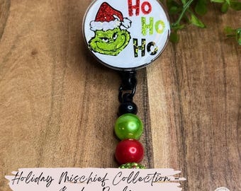 May include: A badge reel featuring the Grinch in a Santa hat with the words "Ho Ho Ho." The reel has a black cord with green and red beads. The text "Holiday Mischief Collection - Badge Reels" is also visible.