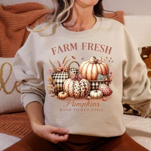 May include: Beige sweatshirt with a fall-themed graphic. The design features pumpkins, a basket, and the text "Farm Fresh Pumpkins Hand Picked Daily" in brown. The pumpkins have various patterns and colors.