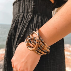 May include: A multi-strand bracelet with a coiled design. The bracelet features alternating bands of gold and dark brown metal. The jewelry is worn on a wrist, with a black outfit in the background. The bracelet is a statement piece.