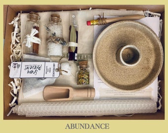 May include: A brown box filled with items for an abundance ritual. The box contains a small white candle, a wooden scoop, a white honeycomb candle, a small bottle of gold glitter, a small bottle of salt, a small bottle of cinnamon sticks, a small bottle of bay leaves, a small bag of herbs, a small tag that says "Abundance Herbs and Flowers", a small tag that says "Stop, Present", a small pencil, and a small ceramic dish.