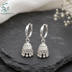 May include: A pair of silver hoop earrings featuring dangling jhumka-style bell charms with intricate embossed patterns and small spherical beads along the bottom edge. The jewelry is branded with the text Barakah Jewelry in the upper left corner.