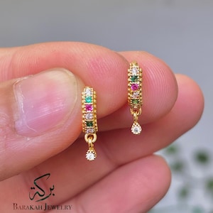 May include: A pair of gold hoop earrings featuring a vertical row of alternating green, pink, and clear stones, with a single clear stone dangling from the bottom of each hoop. The text Barakah Jewelry is printed in black at the bottom left.