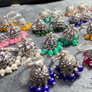 May include: An assortment of silver-toned rings with decorative bell-shaped pendants. The pendants feature colorful beaded accents in shades of pink, yellow, green, blue, and white. The rings are displayed on a gray surface. The image includes the text "Barakah Jewelry".