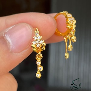 May include: A pair of gold-colored earrings with a floral design and dangling accents. The earrings are adorned with small, clear gemstones. One earring is held up, showcasing the intricate details and craftsmanship.