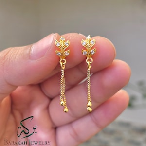 May include: A pair of gold-colored leaf-shaped earrings. Each earring features a leaf design with small, clear gemstones and a dangling chain with small gold beads. The earrings are from Barakah Jewelry.
